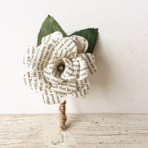 May include: A white paper flower made from book pages with a pearl accent. The flower is attached to a brown twine stem.