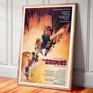 Puede incluir: Un póster enmarcado de la película "Los Goonies" muestra a un grupo de niños en una escena aventurera. El póster tiene un marco de madera marrón. El título "Los Goonies" se muestra de forma destacada, con los nombres del director y los actores.