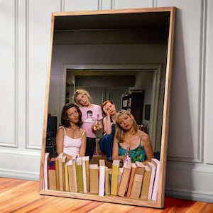 Op de afbeelding: Een ingelijste print met vier vrouwen die in een spiegel worden weerspiegeld. De spiegel is omlijst met licht hout en staat tegen een witte muur. De vrouwen bevinden zich in een kamer met boeken en een televisie. De vrouwen dragen vrijetijdskleding.
