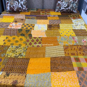 May include: A patchwork quilt with a variety of yellow, orange, and brown fabrics. The quilt is made of squares and rectangles of different patterns and textures. Two decorative pillows with floral patterns are on top of the quilt. The text "VaibhavLaxmiExports" is visible.