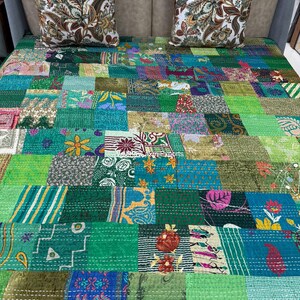 Op de afbeelding: Een patchwork quilt met een levendige mix van groene, blauwe en andere kleurrijke stoffen vierkanten. Twee decoratieve kussens met een bloemenpatroon liggen erop. Een uniek, handgemaakt woondecoratie-item.