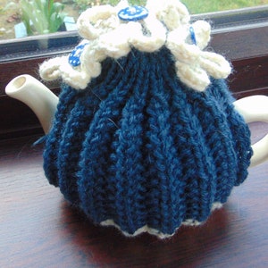 May include: A blue knitted teapot cosy with a white crocheted flower on top. The cosy has a white crocheted trim around the bottom.