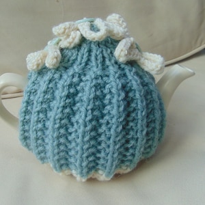 May include: A teal blue knitted teapot cosy with white crocheted flowers. The cosy is designed to fit a standard sized teapot.