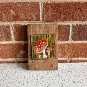 May include: A rectangular wooden plaque with a ceramic tile featuring a red and white mushroom design. The tile is set against a green and brown background, with a rustic, natural aesthetic. The wooden frame adds a touch of warmth.