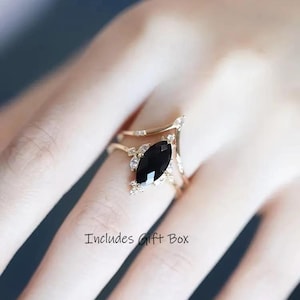 May include: A gold-toned ring featuring a large, black, marquise-cut gemstone as the focal point. The ring has a delicate, ornate design with smaller, clear accent stones. The text "Includes Gift Box" is visible.