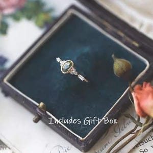 May include: A gold ring with an oval green gemstone and small clear stones, presented in a dark blue gift box. The ring is on a dark blue velvet surface. The text "Includes Gift Box" is visible.