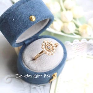 May include: A gold ring with a diamond center stone and a floral design, set in a blue velvet gift box. The ring features a diamond-shaped center surrounded by small pearls. The gift box is open, revealing the ring. The text "Includes Gift Box" is visible.