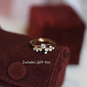 Puede incluir: Un anillo dorado con un grupo de pequeñas piedras preciosas transparentes. El anillo está sobre una superficie de terciopelo burdeos. El texto "Includes Gift Box" es visible.