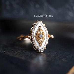 May include: A gold ring with an oval, white centre featuring a filigree design and a central diamond. The ring is decorated with small, clear gemstones around the oval. The text "Includes Gift Box" is visible.