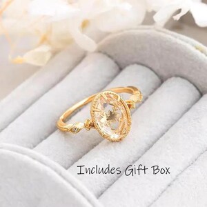 Puede incluir: Un anillo de oro con una piedra preciosa ovalada de color claro con un diseño floral. El anillo tiene una banda delicada con detalles decorativos y se presenta en una caja de regalo blanca. El texto "Includes Gift Box" es visible.