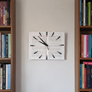 May include: A unique wall clock made from an open book, with pencil-shaped markers indicating the hours. The clock hands are black, and the pencils are green and black. The clock is mounted on a white wall, flanked by two bookshelves.