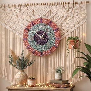 May include: A circular wall clock featuring a mosaic of crushed red, turquoise, and purple stones, with gold-coloured hands. It is mounted on a large, cream-coloured macrame wall hanging with intricate knotted patterns and long fringe.