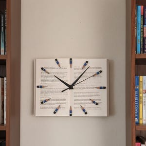 Puede incluir: Un reloj de pared único hecho de un libro, con marcadores en forma de lápiz para las horas. Las manecillas del reloj son negras y los lápices son azules y amarillos. El reloj está montado en una pared beige.