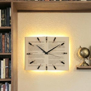 May include: A unique wall clock designed to resemble an open book, with black clock hands and pencil-shaped markers. The clock face features printed text and is illuminated with a warm, soft light. A creative home decor item.
