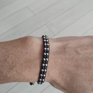 Op de afbeelding: Een armband met twee rijen kralen, bestaande uit afwisselend zilverkleurige metalen kralen en donkere, iriserende paarse kralen geweven op een zwarte koordbasis. De armband heeft een gestructureerde, parallelle rangschikking van kralen.