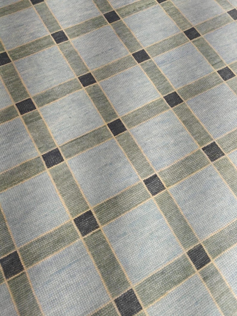 May include: A close-up view of a patterned rug. The rug features a grid design with light blue squares, bordered by olive green and tan lines. Dark blue squares are at the intersections of the grid lines. The rug has a textured appearance.