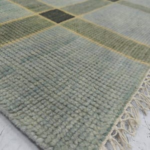 May include: A close-up of a rectangular rug with a plaid pattern. The rug features a color palette of light blue, green, and black squares, with yellow accent lines. The rug has a textured surface and a fringe edge.