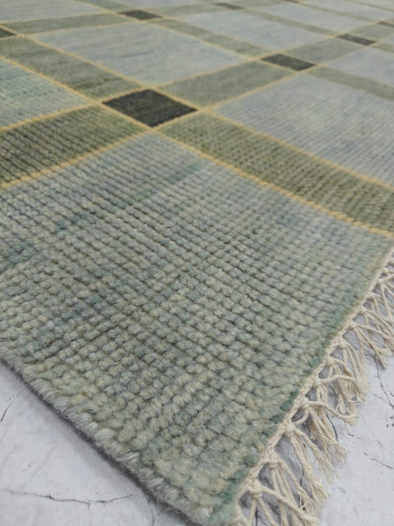 May include: A close-up of a rectangular rug with a plaid pattern. The rug features a color palette of light blue, green, and black squares, with yellow accent lines. The rug has a textured surface and a fringe edge.