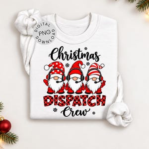 Op de afbeelding: Een wit sweatshirt met lange mouwen en een afbeelding van drie kabouters met rood-witte mutsen en headsets. De tekst luidt Christmas DISPATCH Crew. De kabouters hebben witte baarden en dragen rode kleding. Het woord DISPATCH is in rood ruitpatroon.