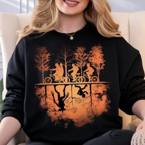 Puede incluir: Sudadera negra con un diseño gráfico naranja y negro. El diseño muestra siluetas de personas en bicicleta sobre una imagen especular de un monstruo y otra figura. También se incluyen siluetas de árboles.