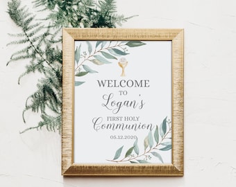 May include: A gold-framed sign with a white background and green foliage design. The sign reads "Welcome to Logan's First Holy Communion 06.12.2020".