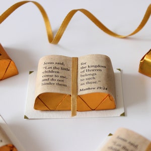 May include: Gold-wrapped Hershey's chocolate nuggets are arranged to look like a small book with a Bible verse printed on the cover. The verse reads "Jesus said, "Let the little children come to me, and do not hinder them" for the kingdom of Heaven belongs to such as these." Matthew 19:14