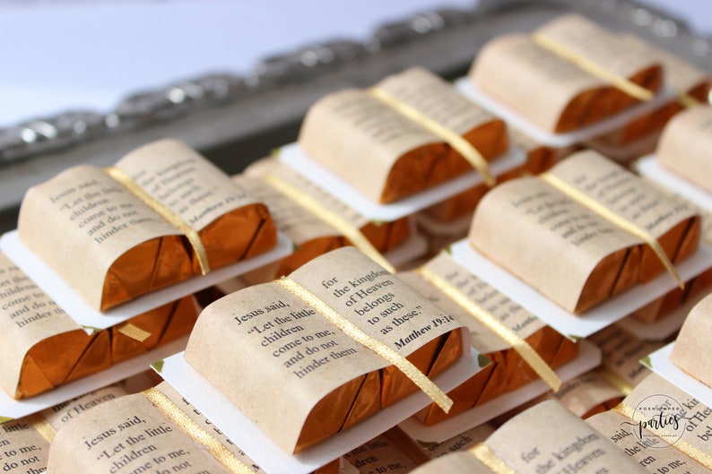 May include: A close-up of several small, wrapped chocolate bars shaped like books. Each book is decorated with a gold ribbon and a printed bible verse. The verse reads "Jesus said, "Let the little children come to me, and do not hinder them." Matthew 19:14"