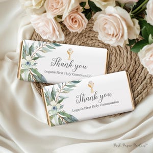 Puede incluir: Dos envoltorios de barra de chocolate blanco con un diseño floral de acuarela y un icono de cáliz dorado. El texto en los envoltorios dice "Gracias Logan's First Holy Communion".
