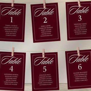 Op de afbeelding: Bordeaux tafelkaarten met witte kalligrafie, genummerd 1-6, zijn aan een touwtje geknipt. Elke kaart bevat namen, ideaal voor bruiloften of evenementen. De kaarten hebben een witte rand en het woord "Table" in elegant schrift.