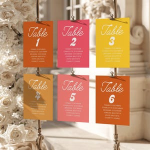 May include: Six rectangular wedding table seating charts in orange, pink, and yellow, suspended by metal clips. Each card features the word Table followed by a number from 1 to 6 in white script, with a list of names printed in white below.