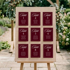 May include: A rectangular beige seating chart board displayed on a wooden easel, featuring nine deep burgundy cards arranged in a grid. Each card displays the word Table followed by a number from 1 to 9 and a list of names in white text.