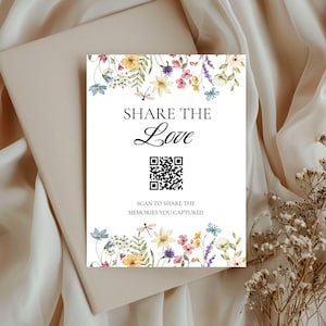May include: A white card featuring the words "SHARE THE Love" and a QR code, adorned with watercolour floral designs in yellow, blue, and purple. The card rests on a beige envelope, against a backdrop of draped fabric.