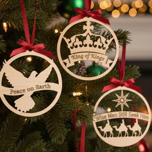 May include: Wooden Christmas ornaments hanging on a decorated tree. The ornaments are round with cut-out designs of a dove, a crown, and three camels. Each ornament has a red ribbon for hanging and features the phrases "Peace on Earth", "King of Kings", and "Wise Men Still Seek Him".