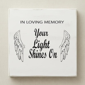 Puede incluir: Un letrero de madera blanco con el texto "IN LOVING MEMORY Your Light Shines On" en escritura negra. El letrero presenta ilustraciones de alas negras a ambos lados del texto. El letrero es cuadrado y tiene un aspecto ligeramente envejecido.