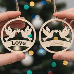 May include: Two circular wooden ornaments featuring a dove design and the word "Love". Each ornament shows two doves facing each other with a heart between them. The ornaments are suspended by string, with a Christmas tree in the background.