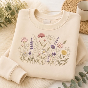 May include: A cream-colored crewneck sweatshirt featuring a detailed embroidered design of various wildflowers in shades of pink, purple, yellow, and white, with green stems and leaves centered on the front of the garment.