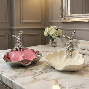 Peut inclure: Deux porte-savons décoratifs. L'un est argenté avec un intérieur rose et des savons roses, surmonté d'une figurine de lapin. L'autre est en forme de coquillage crème avec une figurine de lapin. Les deux sont sur un comptoir en marbre.