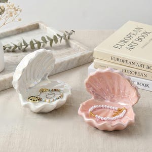 May include: Two ceramic seashell-shaped trinket dishes. One is white, the other is pink. The white dish holds a gold ring and a beaded bracelet. The pink dish holds a pearl bracelet. "European Art Book" is visible in the background.