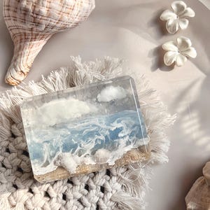 May include: A rectangular soap bar with a beach scene design, featuring blue waves, white clouds, and seashells. The soap rests on a macrame coaster, with a seashell and white flower accents.