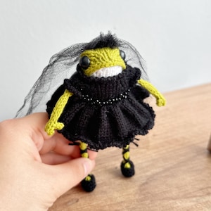 May include: A handmade, knitted frog doll wearing a black dress, veil, and beaded necklace. The frog has a green head and yellow arms and legs. The doll's feet are black, and the frog is wearing black shoes.