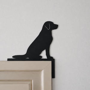 Puede incluir: Una silueta de metal negro mate de un perro sentado, diseñada para descansar sobre la esquina superior del marco de una puerta. El perro se muestra de perfil, mirando hacia la derecha, con el contorno de una oreja visible. La pieza tiene una base plana.