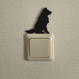 Op de afbeelding: Een zwart silhouet van een zittende hond is direct boven een vierkante, gebroken witte lichtschakelaar geplaatst. De hondenfiguur is uitgevoerd in een effen matzwarte afwerking en toont het profiel van een hond met een pluizige staart en spitse oren.