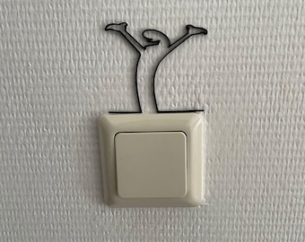 May include: A light switch cover featuring a black, thin, wire-like decorative topper shaped as a stylized figure with arms raised upward. The figure is positioned directly above the square, off-white plastic light switch plate on the wall.