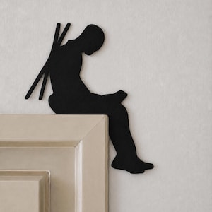 Può includere: Una sagoma nera di una persona seduta sul bordo di una cornice, che tiene un piccolo oggetto rettangolare in grembo. Due lunghe aste sottili e dritte si estendono diagonalmente verso l'alto da dietro la schiena della persona.