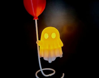May include: A glowing yellow ghost figurine with two circular eyes, holding a bright red balloon attached to a white, spiraled base. The ghost emits a soft light, contrasting against the dark background, creating a distinct silhouette.
