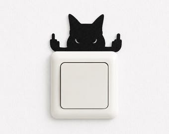 May include: A white square light switch cover featuring a black silhouette of a cat head positioned above it. The cat figure has pointed ears, narrow eyes, and two arms extended outward with the middle fingers raised in a rude gesture.