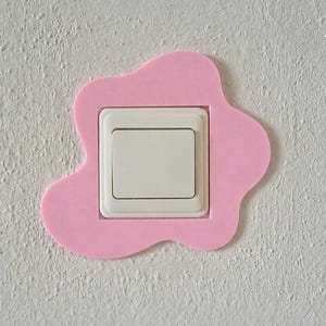 Op de afbeelding: Een witte vierkante lichtschakelaar aan een muur, omringd door een roze, onregelmatig gevormd, vlekachtig decoratief frame. Het frame heeft gladde, afgeronde randen en een plat oppervlak, wat een kleurrijke rand rond de schakelaar vormt.