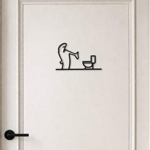 Pode incluir: Arte de parede em metal preto que retrata uma pessoa usando um vaso sanitário. O design minimalista apresenta um contorno simples de uma figura e um vaso sanitário, montado em uma porta branca. A porta tem uma maçaneta preta e uma moldura branca.