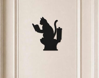 May include: A black silhouette wall decal featuring a cat sitting on a toilet while reading a book. The cat is depicted in profile, holding an open book with its front paws, with its tail curved upward. The design is flat, solid black, and matte.