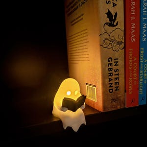 May include: A small, glowing white ghost figurine reading a black book, positioned on a shelf next to three books titled In Steen Gebrand by Rebecca Yarros, A Court of Thorns and Roses by Sarah J. Maas, and A Court of Frost and Starlight by Sarah J. Maas.